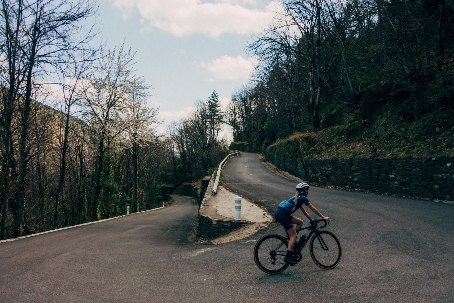 Radfahrerin fährt auf kurviger Straße durch bewaldete Hügel, Bäume ohne Blätter und bewölkter Himmel im Hintergrund.