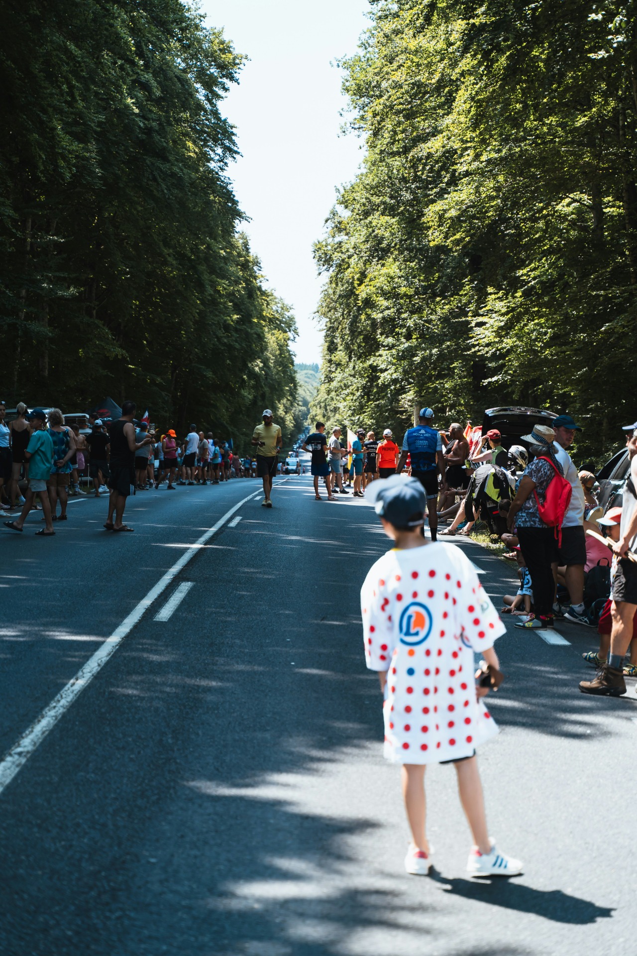 Menschen stehen am Straßenrand einer Tour de France Etappe auf einer von Bäumen gesäumten Allee.