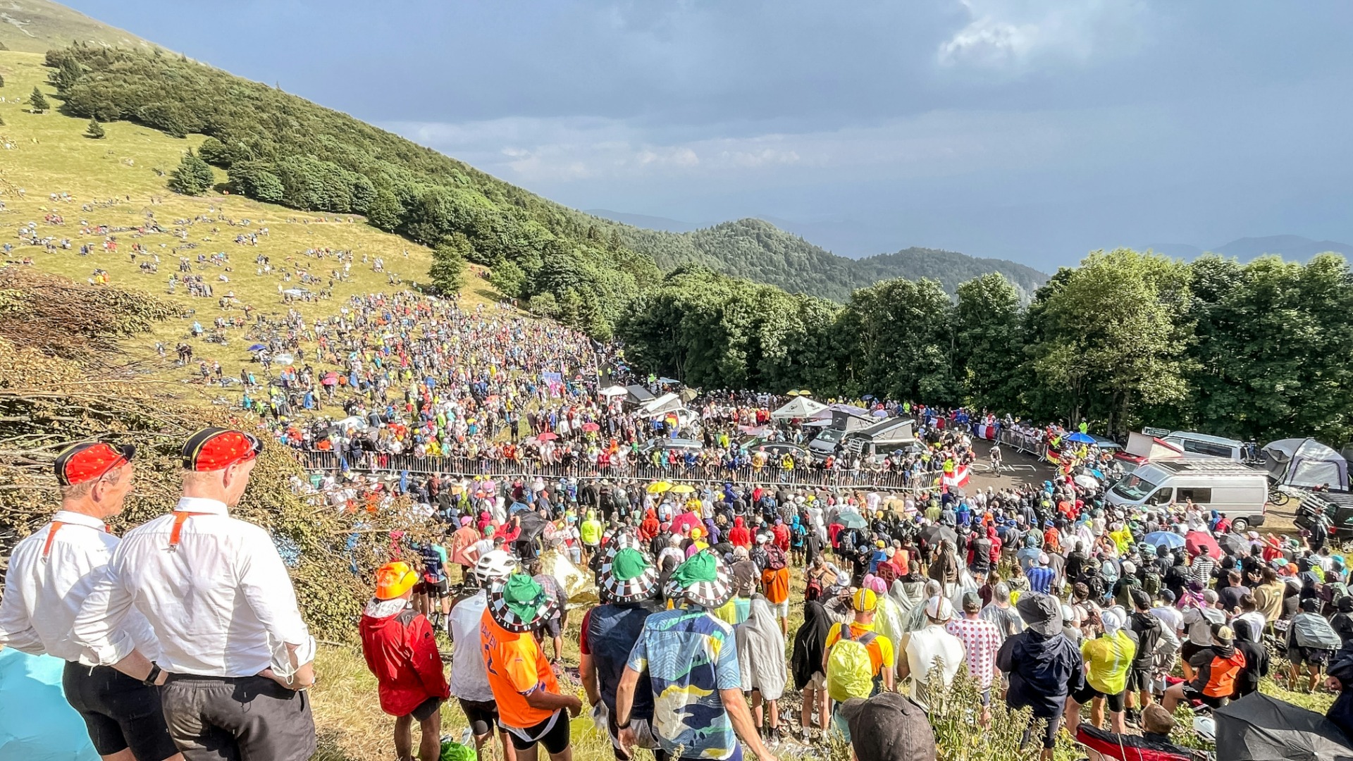 Große Menschenmenge bei der Tour de France, Zuschauer stehen auf steilem Hang, Fahrzeuge und Zelte am Straßenrand.