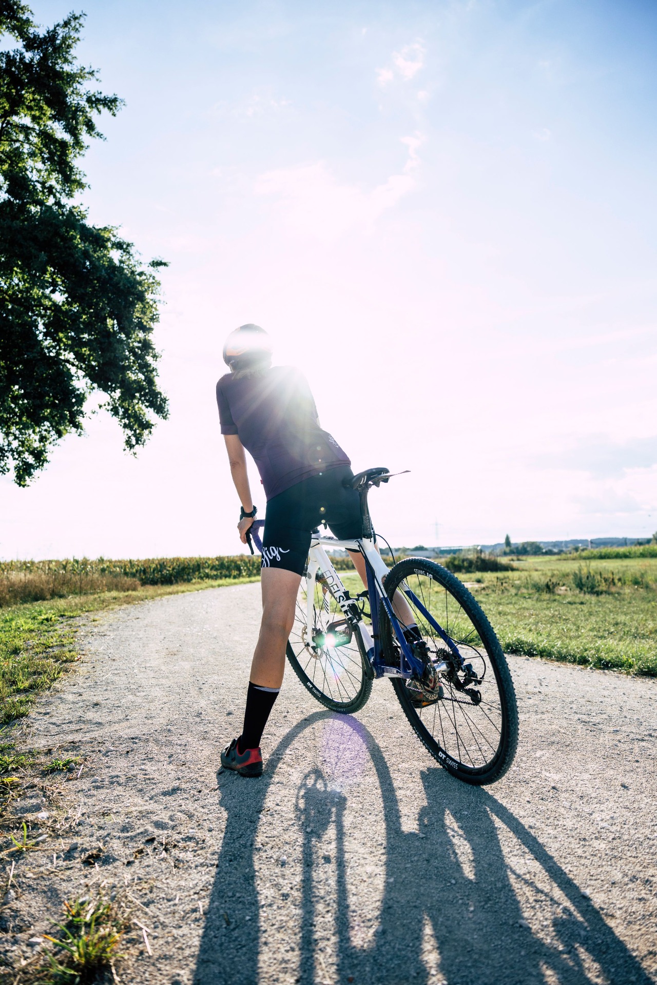 Radfahrer steht auf Feldweg, trägt Helm und hält Fahrrad bei Sonnenlicht, umgeben von Wiesen und Bäumen.