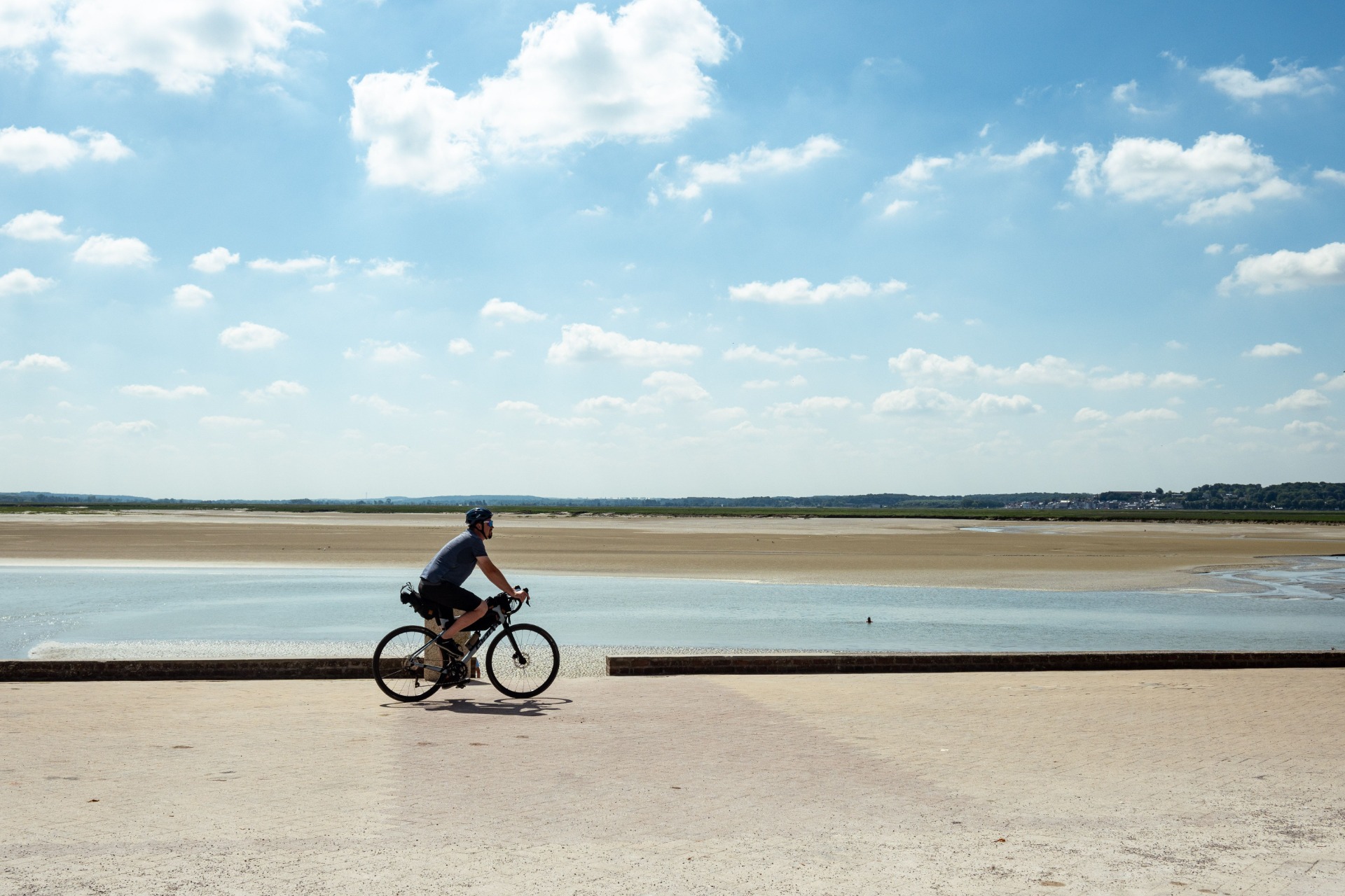 Radfahrer fährt am Ufer entlang, im Hintergrund breite Sandflächen, Wasser und weitläufiger Himmel mit Wolken.